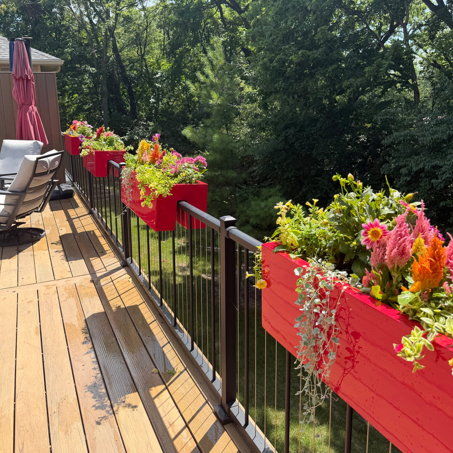 A Des Moines residential deck with gloss red rectangular railing planters filled with green and pink seasonal plants, overlooking dense forest.