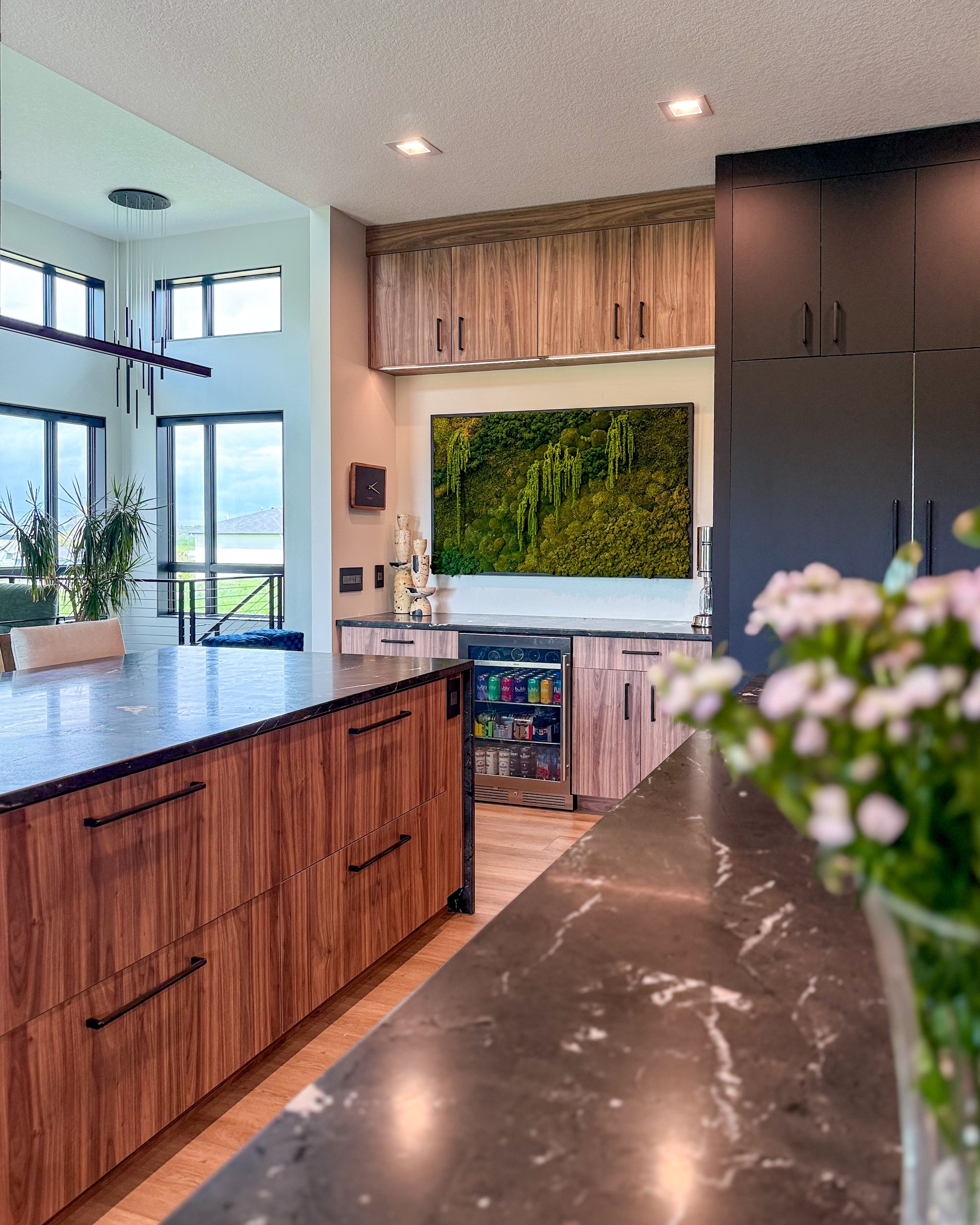 Modern kitchen featuring a custom framed preserved moss wall art installation above a beverage station, blending reindeer and mood moss textures for natural, maintenance-free greenery. Surrounded by warm wood cabinetry, matte black finishes, and panoramic windows, this luxury moss wall brings biophilic interior design into a residential space.