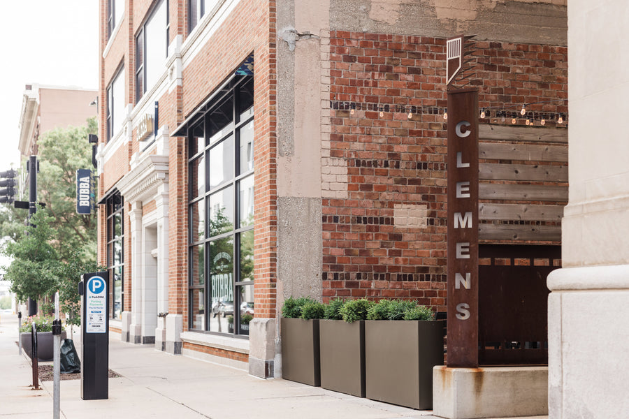 Three rectangular semi-custom fiberglass planters in matte bronze with boxwood dividing the sidewalk and the outdoor patio entrance of Bubba in downtown Des Moines.